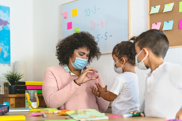 Teacher cleaning hands to student children with sanitizer gel while wearing face mask in preschool classroom during corona virus pandemic - Healthcare and education concept - obrazy, fototapety, plakaty