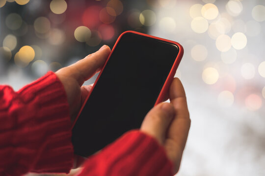 Cropped of woman's hands in red sweater holding smartphone with blank screen of gadget at Christmas time. Digital mobile phone with copy space area