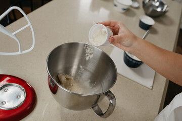 A close photo of the hand of a young woman who is putting an almond flour from the cup into the stainless steel bowl of the stand mixer.