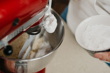 A close photo of the spoonful and a cup of powdered sugar above the stainless steel bowl of the red stand mixer which is mixing shortcrust pastry.