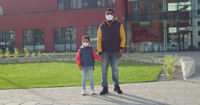 African American Male Parent In Mask Standing Outdoors At School With Cute Little Son Pupil Before Classes Young Father Bringing Junior Student To School In Quarantine Education Concept Back To School