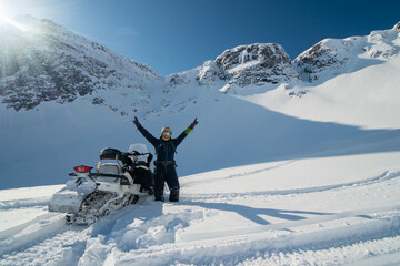 A man next to a snowmobile in the mountains. A lot of snow, blue winter sky and the rays of the...