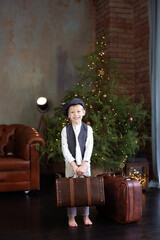 Little boy with suitcase in hand in Cozy christmas living room. Smiling Little blond boy in white shirt with vest and in cap sit by Christmas tree with old suitcases. 