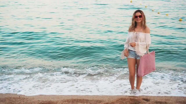 Happy beautiful woman tourist stands and dreams by sea coast. Boho style clothing, red bag, vintage sunglasses. Turquoise water waves. Happy girl travel in Spain. Costa Brava beach, city Lloret de Mar
