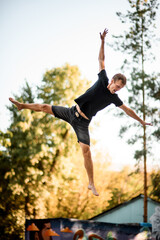 Athletic man skillfully jumps on trampoline against the backdrop of green trees and sky.