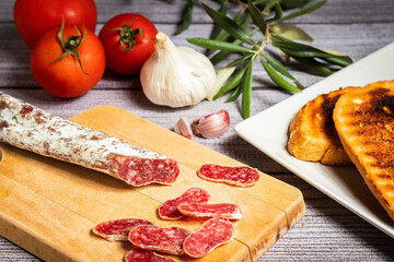 Trozitos de fuet encima de tabla rústica marrón claro con ajos y tomates en un fondo gris rústico claro
