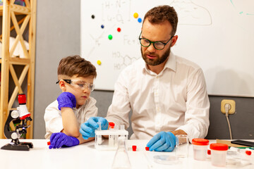 dad and son the child stayed at home conduct experiments on a white table, behind them on the wall is a white Board magnetic-marker
