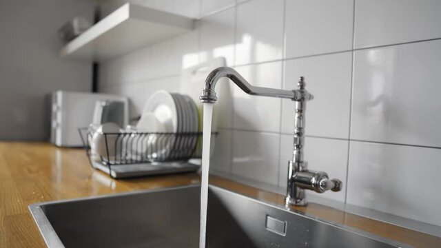 Water flowing out of a kitchen stainless steel tap into the sink. Wasting water by leaving a chrome faucet tap running. Water misuse in domestic duties and activities. Overusing household water.