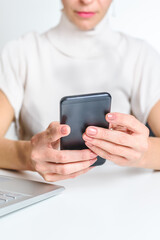 hands of an unrecognizable young caucasian woman using a smart phone near a laptop on a white desk