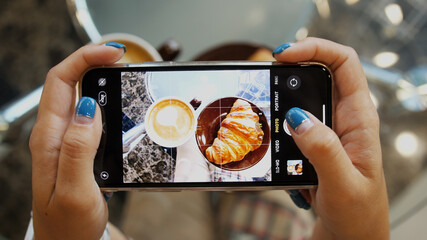Close up of woman holds modern smartphone and taking pictures of her morning breakfast coffee and tasty croissant buns to share photos on social media resources