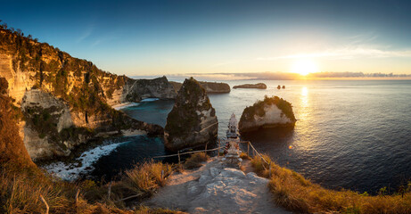 Fototapeta na wymiar Panorama sunrise over steep cliffs and crystal clear ocean with small temple in foreground at Thousand Islands viewpoint, Nusa Penida, Bali, Indonesia