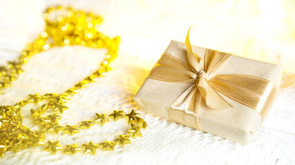 Box with Christmas gift in light paper is tied with a gold ribbon on white knitted background with decor. Golden garland of stars. New Year. Congratulations, surprise. space for text