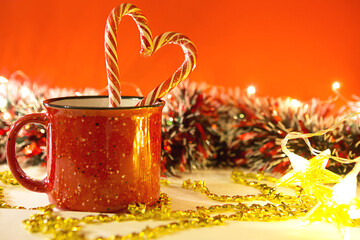 Red mug with striped candy canes in the shape of a heart on a red Christmas background with lights of garlands in bokeh. Copy space. New Year.