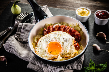 Fried sausages with egg and onion in frying pan on wooden table
