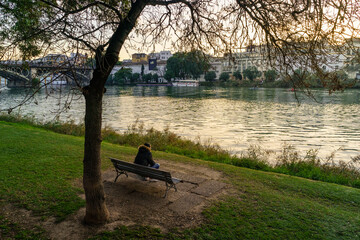 Chico de espaldas sentado en un banco al lado del río Guadalquivir mirando al frente.