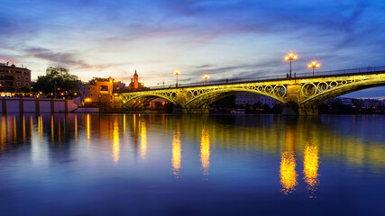 Puente de Triana en Río Guadalquir en Sevilla, Andalucía. Puesta de sol con reflejos en el agua y luces de farolas. 
