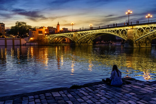 Chica de espaldas sentada en el suelo mirando al r&iacute;o Guadalquivir y al puente de triana al atardecer.