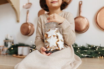 Toddler girl wearing beige dress holding glass jar with christmas cookies