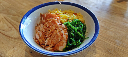 Japanese rice bowl filled with salmon, sliced sweet egg slice and seaweeds on wooden table.