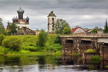 Old Church of St. Paraskeva on the opposite bank covered with green grass and wooden bridge over the river against cloudy sky in Izmailovskaya village, Arkhangelsk region, Russia.