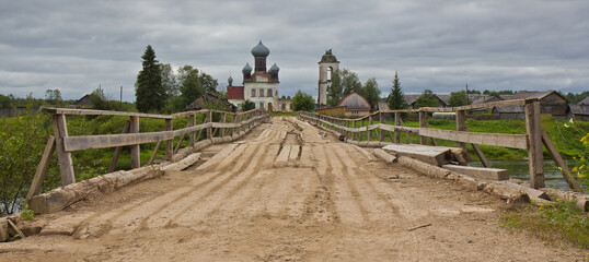 A wooden bridge over the river and old Church of St. Paraskeva on the opposite bank against cloudy sky in summer in Izmailovskaya village, Arkhangelsk region, Russia.