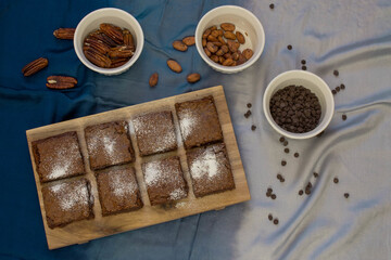 Chocolate brownies on a wooden board with chocolate chips, almonds, and pecans on a white bowl