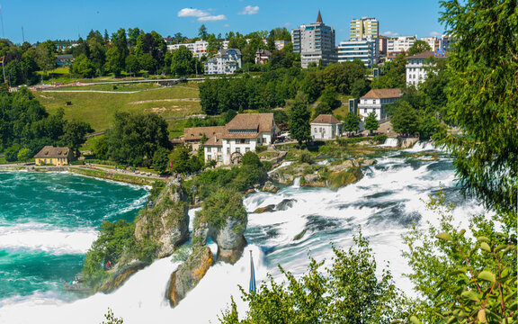 Panorama des cascades du Rhin ou Rheinfall en Suisse par une journ&eacute;e ensoleill&eacute;e avec un ciel bleu avec les rochers et un village et les embruns du fleuve.