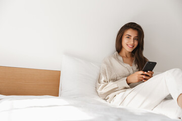 Technology, communication and people concept - happy young woman with smartphone texting message in bed at home. Girl using mobile phone and relaxing in bedroom