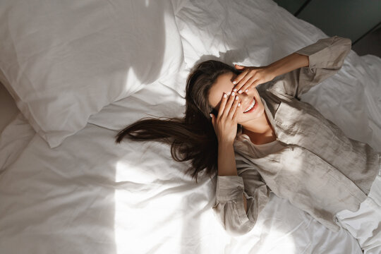 High-angle shot of young woman lying in bed, rubbing her eyes and smiling. Concept of relaxing morning and lifestyle