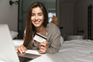 Image of young woman with credit card and laptop shopping in internet, lying in bed and smiling at camera
