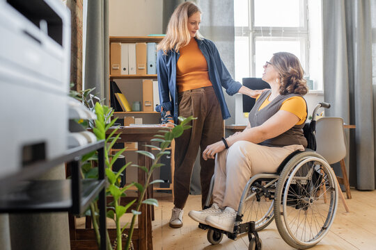 Young blond woman in casualwear looking at her disable friend in wheelchair