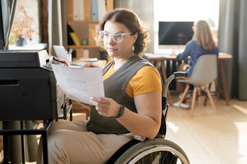 Young disable office worker looking at paper while sitting by xerox machine