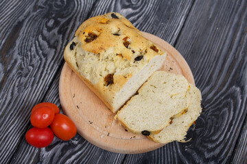 Focaccia bread with olives and sun-dried tomatoes. Several red tomatoes are nearby. On pine boards.