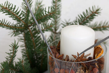 A candle in a glass candlestick. The candlestick is filled with pine cones and spices. Nearby are fir branches