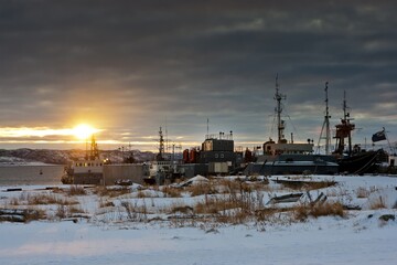 Sunset and cloudy sky over a dock full of moored ships in winter and the ground covered with snow and faded grass. Polyarny, Russia.