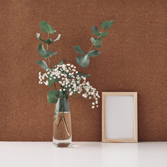 Wooden frame and vase with white flowers and eucalyptus branches, cork background. Mock up, copy space. Folk.