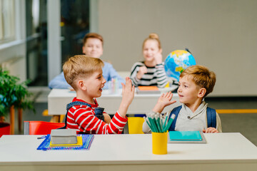 Fototapeta premium Two handsome blond boy greeting at morning, giving high five, sitting at workplace in classroom. Frindship, elementary school, real people model concept.