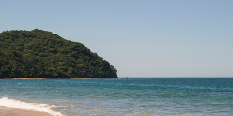 Beautiful landscape of a beach in the mexican pacific coast in a sunny day with a blue and clear sky, trees, palms and green hills, some waves and sand in the beach