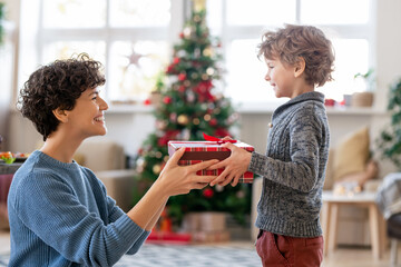 Young laughing mother passing Christmas gift to her adorable little son at home
