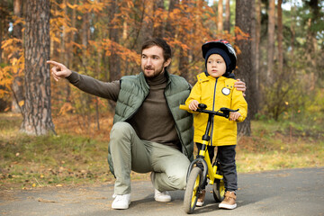 Happy young bearded man pointing forwards while squatting by his cute little son