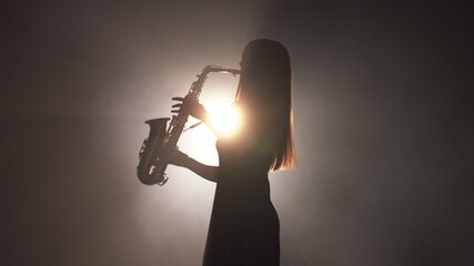 Young beautiful girl in a dark dress plays on a golden shiny saxophone on stage. Dark studio with...