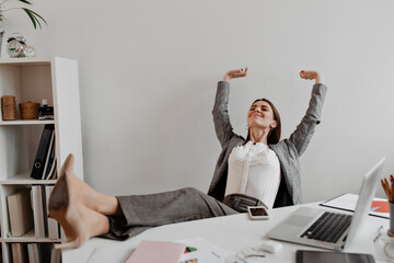 Business lady satisfied with working result with pleasure threw her legs to white desktop