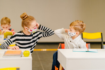 Two pupils kid in protextive mask at classroom greeting each other with elbow bumps while...