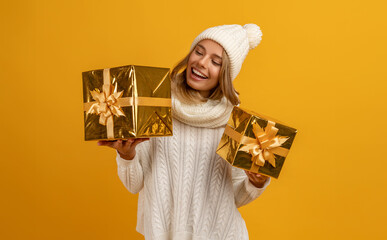 young smiling happy pretty woman holding golden present boxes celebrating new year, christmas gifts, wearing white knitted sweater, scarf and hat, winter fashion trend, posing on yellow background