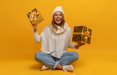 young smiling happy pretty woman sitting with golden present boxes celebrating new year, christmas gifts, wearing white knitted sweater, scarf and hat, winter fashion trend, on yellow background