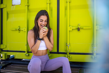 Portrait of happy smiling young beautiful woman in fitness wear with apple, outdoors. Sporty girl relaxing after exercising. Beautiful fit young woman eating apple outside while smiling