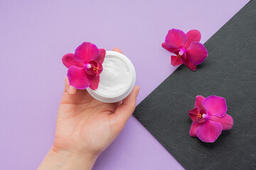 Obraz na płótnie Canvas Background with hand holding soft white cream and fragile magenta colored orchids.Moisturizing facial cream in a jar and blooming orchid flowers on purple and black background, skin care cosmetics.