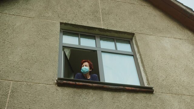 Old woman wearing covid 19 medical protective mask looking outside her window during lockdown.