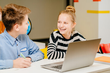 Portrait of happy laughing diligent pupil looking at her classmate at lesson. Children of elementary school sitting with laptop computer at workplace in classroom.