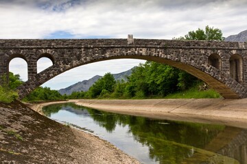 Fototapeta na wymiar A stone bridge over the artificial channel with concrete embankment surrounded with green bushes and mountains in the background in Bosnia and Herzegovina.